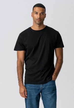 Mann med kort hår som har på seg en enkel svart t-skjorte og blå jeans, stående med den ene hånden i lommen mot en ensfarget bakgrunn.