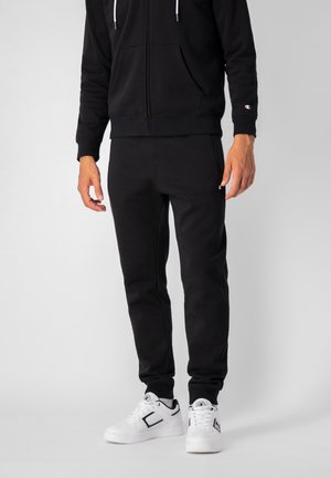 Felpa nera con zip e pantaloni della tuta abbinati, entrambi in un tessuto morbido, con tasche laterali e lacci bianchi, abbinati a sneakers bianche.