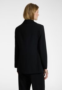 Una donna con capelli biondi corti indossa un blazer nero aderente e pantaloni neri, in piedi di spalle su uno sfondo bianco semplice.