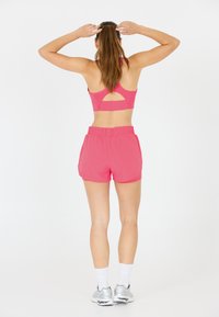 Vaaleanpunainen urheiluliivi, jossa on leikattu yksityiskohta, yhdistettynä yhteensopiviin vaaleanpunaisiin shortseihin. Mallilla on valkoiset sukat ja hopeiset juoksukengät.