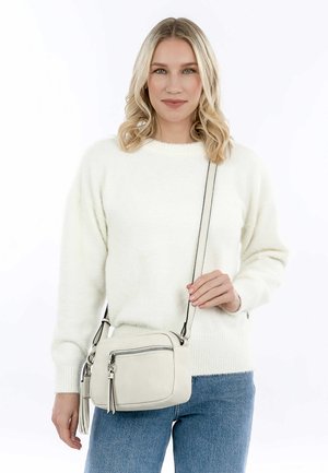 Beige Umhängetasche aus strukturiertem Leder, mit Reißverschluss, Vorderfach und dekorativen Quasten, getragen über einem weißen Pullover.