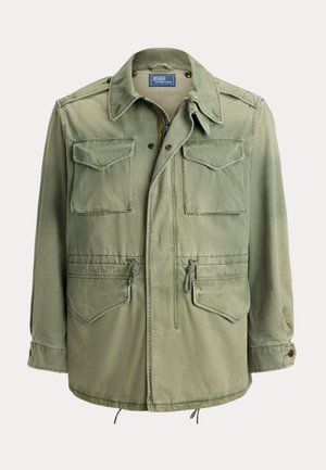 Mand iført solbriller, grøn militærinspireret jakke, mønstret sweater, hvide bukser og brune loafers, der holder en stor brun lædertaske.