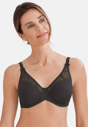 Frau trägt einen schwarzen Bügel-BH mit Spitze, lächelt leicht und schaut seitlich vor einem schlichten hellen Hintergrund.