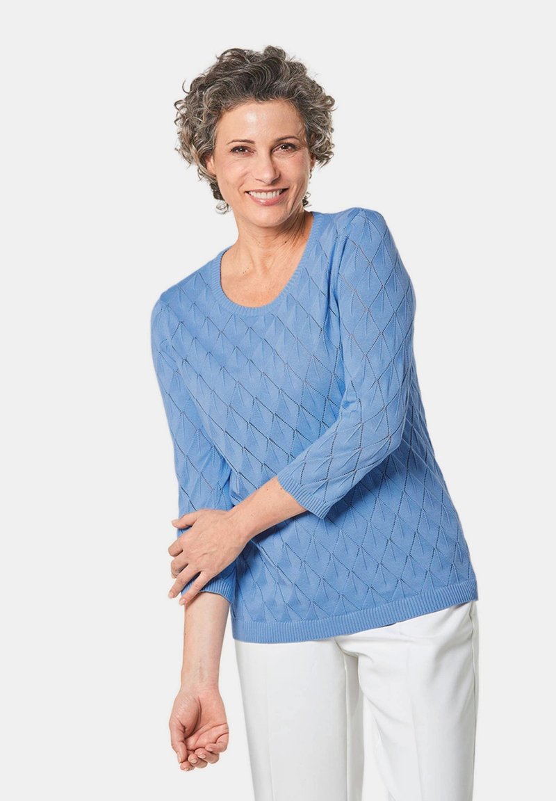 Hellblauer Pullover mit Rautenmuster, geripptem Hals- und Armabschlüssen, Dreiviertelärmeln, kombiniert mit weißen Hosen. Texturiertes Strickmaterial.
