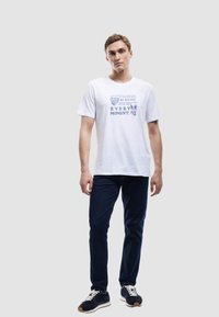 Camiseta de algodón blanca con texto y gráficos en color azul. Combinada con jeans oscuros y zapatillas negras, sobre un fondo neutro.