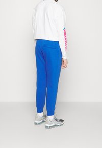 Siniset verkkarit, joissa takatasku, yhdistettynä valkoiseen pitkähihaseen paitaan, jossa vaaleanpunaiset ja siniset raidat hihassa. Harmaat lenkkarit, joissa kirkkaat pohjat.