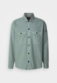 Chemise longue à manches longues de couleur vert clair avec deux poches poitrine et des boutons noirs, dotée d'un col et d'un écusson sur la manche gauche.