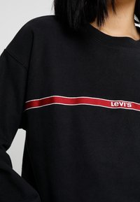 Felpa nera in cotone con scollo rotondo, caratterizzata da una striscia orizzontale rossa e bianca con il marchio "Levi's" in rosso.