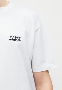 Balta īsroku T-krekls ar melnu tekstu "the new originals", izšūts uz kreisā krūšu apgabala, ko valkā persona uz gaiša fona.