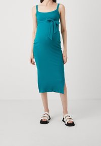 Türkisfarbiges, geripptes Umstandskleid mit taillierter Silhouette, quadratischem Ausschnitt, seitlichem Schlitz und verstellbarem Bindegürtel in der Taille, kombiniert mit schwarz-weißen Sandalen.