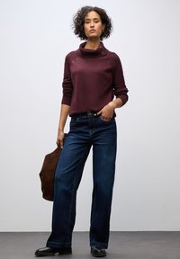 Bordeauxfarbener Strickpullover mit hohem Kragen und Knopfdetails, kombiniert mit dunkelblauen weiten Jeans und schwarzen Ankle Boots. Braune Wildledertasche in der Hand.