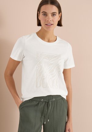Junge Frau trägt ein weißes T-Shirt mit abstrakten Linien und olivgrünen Kordelzughosen, steht vor einem beigen Hintergrund.