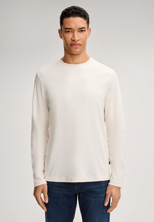 Langärmliges cremefarbenes T-Shirt aus weichem Stoff, mit rundem Ausschnitt und kleinem Logo-Detail auf der Vorderseite, kombiniert mit blauen Jeans.