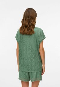 Conjunto de encaje verde que incluye una blusa de corte holgado con mangas tipo capucha y pantalones cortos a juego. Destaca un patrón perforado notable y detalles en el dobladillo con puntas.