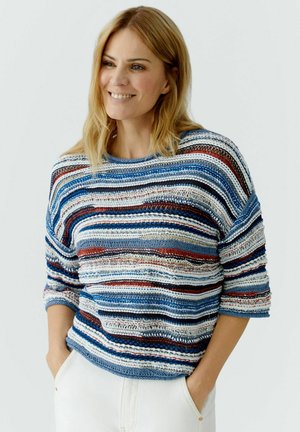 Vrouw lacht met handen in haar zakken, draagt een losse, gestreepte gebreide trui in blauw, wit, rood en zwart, met een witte broek tegen een effen achtergrond.