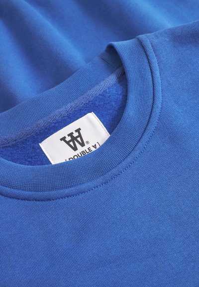 Camiseta de algodón azul con cuello redondo, costuras visibles y una etiqueta que muestra "DOUBLE V". Textura suave sin patrones.