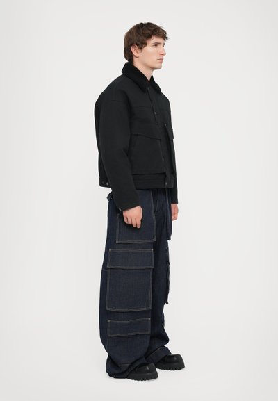 Black jacket with a fur-lined collar, paired with loose-fitting blue denim cargo pants featuring multiple pockets and contrasting stitching.