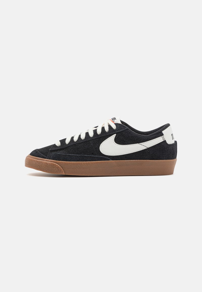Fekete velúr sportcipők fehér Nike logóval, krémszínű fűzővel és barna gumitalppal. Alacsony szárú kialakítás textúrált anyagból.
