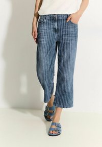 Pantalones vaqueros cortos con un patrón de rayas verticales en un tono azul claro. Cuentan con un cierre de botón frontal y bolsillos laterales. Llevados con sandalias azules.