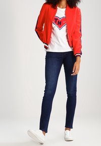 Röd bomberjacka med blå randiga detaljer, vit grafisk t-shirt med ett hjärtmotiv, mörka tajta jeans och vita sneakers.