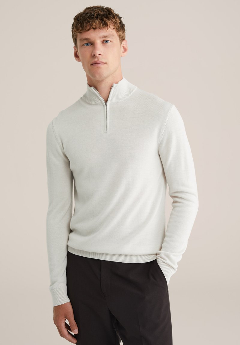 Hellgrauer Strickpullover mit einem Zip-Kragen, langen Ärmeln und gerippten Bündchen, kombiniert mit dunklen Hosen für einen Kontrast.