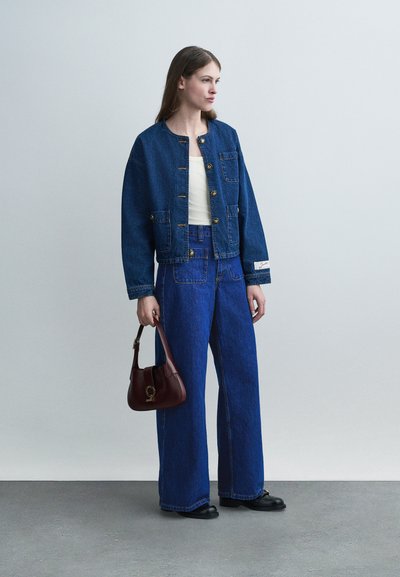 Γυναίκα φοράει σκούρο μπλε τζιν μπουφάν με κουμπιά και παντελόνι με φαρδιά πόδια, κρατώντας μια μικρή μπορντό τσάντα, στέκεται σε γκρι δάπεδο μπροστά σε γκρι τοίχο.