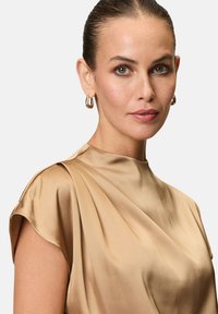 Top in satin a maniche corte di colore beige con morbide drappeggi e un collo alto. Presenta orecchini a cerchio in oro minimalisti.