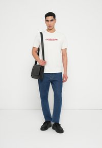 Λευκό βαμβακερό t-shirt με κόκκινο λογότυπο "CALVIN KLEIN", συνδυασμένο με σκούρα μπλε τζιν παντελόνι και μαύρη δερμάτινη τσάντα ώμου. Οι μαύρες αθλητικές παπούτσια ολοκληρώνουν την εμφάνιση.