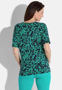 Gemustertes Kurzarmoberteil mit schwarzem Hintergrund und grünem Blattdesign. Weicher Stoff, entspannter Schnitt. Von hinten betrachtet, kombiniert mit türkisfarbenen Hosen.