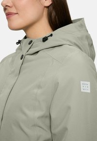 Femme en veste à capuche gris clair avec des boutons pression noirs et un patch logo sur la manche supérieure, faisant face à gauche.