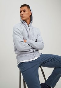 Grå zip-hoodie med snor til hætten, ribbede manchetter og kant, båret over en hvid skjorte, parret med mørkeblå bukser.