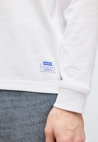 Κοντινό πλάνο ατόμου που φοράει λευκό πουκάμισο μακρυμάνικο με μπλε ετικέτα "HUGO" κοντά στο τελείωμα και μπλε τζιν παντελόνι.