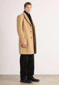 Camel wool overcoat with a tailored fit, notched lapels, and four buttons. Paired with a black turtleneck and black trousers.