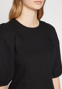 Czarna sukienka z pulowerem, z krótkimi bufkami, okrągłym dekoltem i gładką fakturą. Posiada przeszycia wzdłuż dekoltu i krawędzi rękawów.