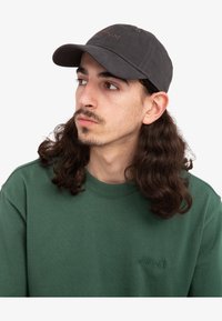 Casquette de baseball grise avec une visière courbée et un logo brodé. Portée avec un sweat-shirt vert arborant un logo ton sur ton subtil sur la poitrine.