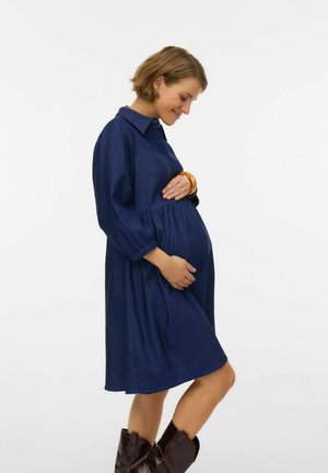 Tumši zilas krāsa, pogātā grūtniecības kleita ar garām piedurknēm, savilkta jostas vietā un plūstoša silueta, kombinēta ar brūnām rietumu stila zābakiem.