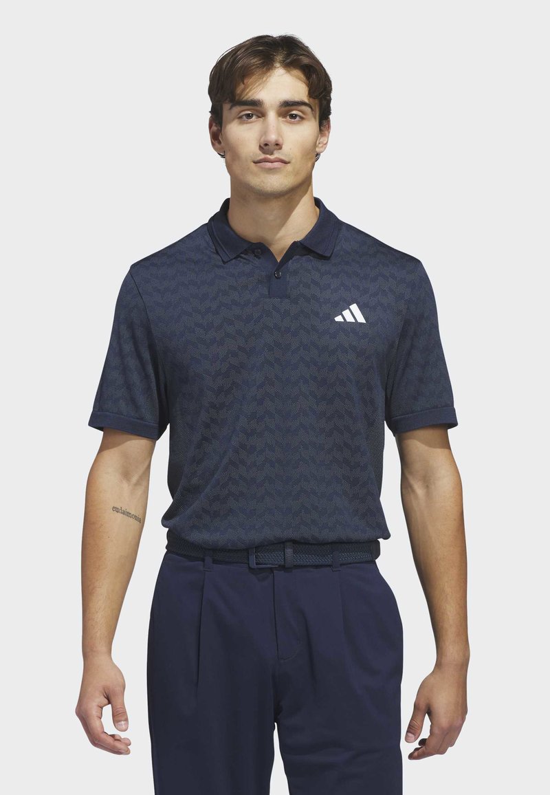 Jonge man staand, gekleed in een marineblauw poloshirt met patroon en wit logo, met bijpassende marineblauwe broek en riem, neutrale achtergrond.