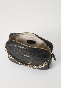 Bolso de piel negra acolchada con una correa de cadena dorada, que cuenta con un cierre de cremallera y un interior beige. Logo mostrado de forma prominente en la parte delantera.