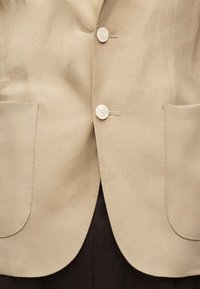 Μπεζ σακάκι με δύο λευκά κουμπιά και δύο μεγάλες μπροστινές τσέπες, φορεμένο πάνω από σκουρόχρωμο παντελόνι.