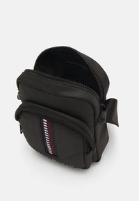 Bolso compacto negro hecho de material texturizado con cierre de cremallera. Presenta un bolsillo frontal con acentos de rayas verticales en rojo, blanco y azul.