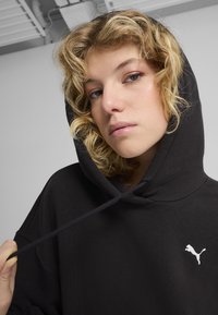 Čierna mikina s kapucňou vyrobená z jemnej tkaniny, ktorá má veľkú kapucňu, šnúrky a biele logo Puma na ľavej strane hrude.