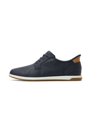 Zapatillas de deporte de cordones en azul marino con un upper de cuero liso, acento en marrón claro en el talón y una suela de goma blanca con textura de agarre.