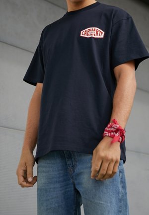 Marineblaues Baumwoll-T-Shirt mit kurzen Ärmeln und rundem Halsausschnitt, mit einem roten und weißen Logo auf der linken Brust. Kombiniert mit blauen Jeans und einem roten Bandana.
