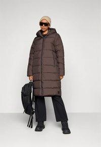 Hosszú, barna steppelt kabát kapucnival és cipzáras záródással, függőleges varrással, oldalzsebekkel, fekete nadrággal és sportcipővel párosítva.