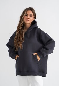 Felpa oversized grigio carbone in tessuto morbido, con tasca a marsupio e polsini a costine. Cappuccio foderato, con un design a vestibilità rilassata.