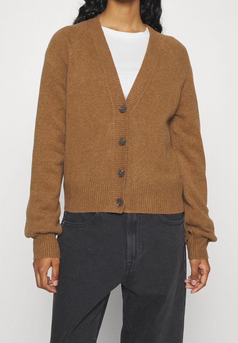 Personne portant un cardigan marron boutonné à col en V par-dessus une chemise blanche et un pantalon sombre ample, debout, les bras détendus le long du corps.