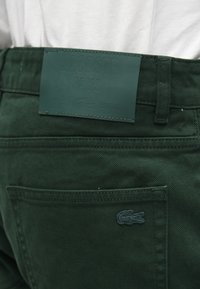 Pantalons en denim vert foncé avec une étiquette en cuir texturé portant "LACOSTE PARIS" embossé. Petit logo brodé sur la poche arrière.