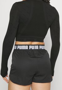 Musta pitkähihainen paita, jossa on teksturoitu kuvio ja napsakka muotoilu, yhdistettynä mustiin shortseihin, joissa on valkoinen Puma-logo vyötäröllä.