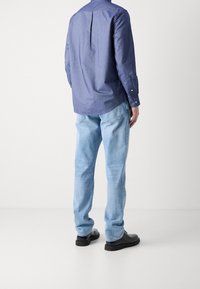 Camicia blu a maniche lunghe con pences posteriori, abbinata a jeans blu chiaro e scarpe nere. La camicia presenta polsini con bottoni e un orlo curvo.