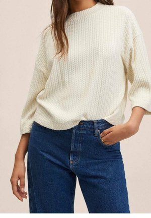 Femme portant un pull en tricot crème à manches trois-quarts et un jean en denim bleu taille haute, une main dans la poche, sur un fond neutre.